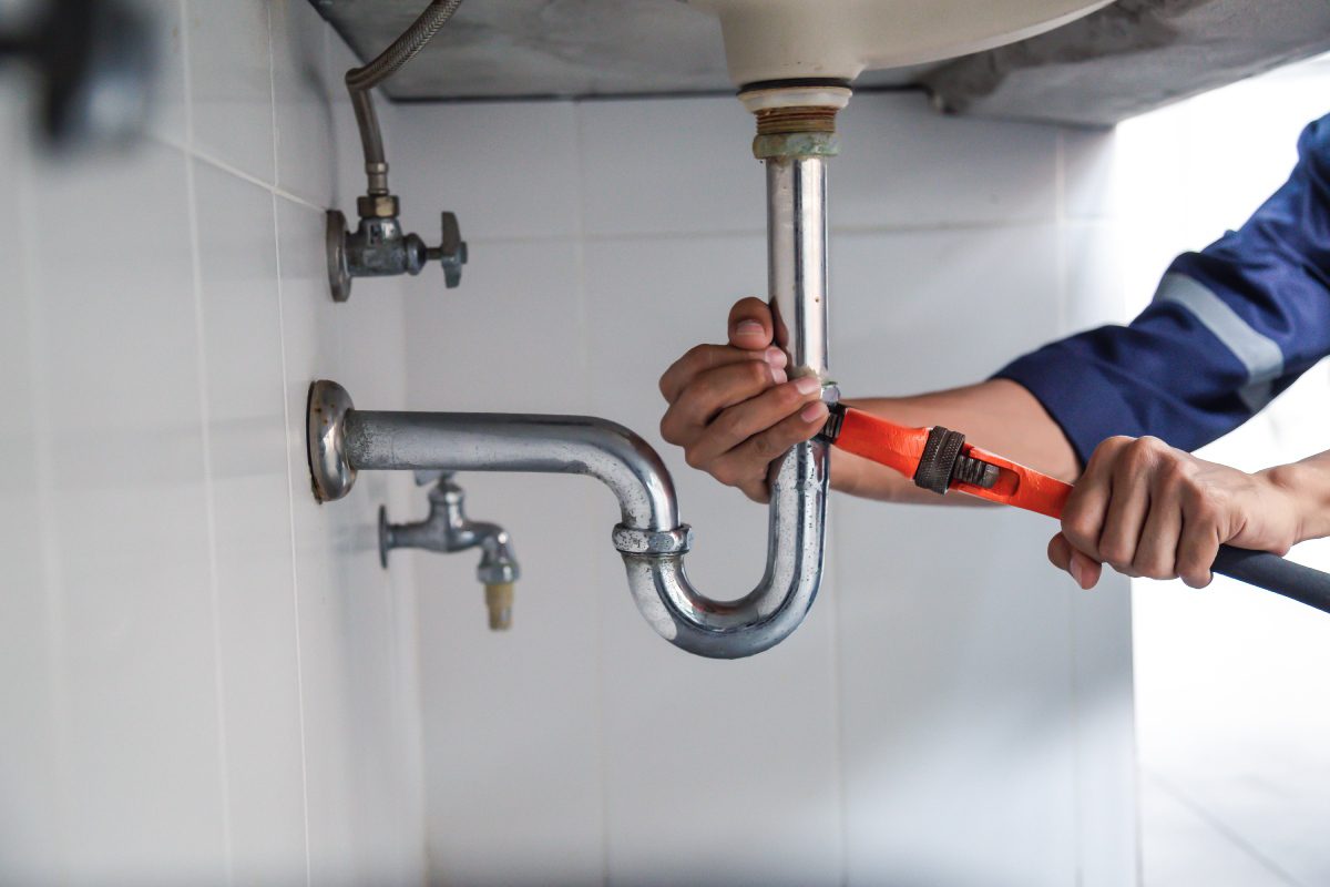 Person fixing a silver pipe under a sink with a pipe wrench.