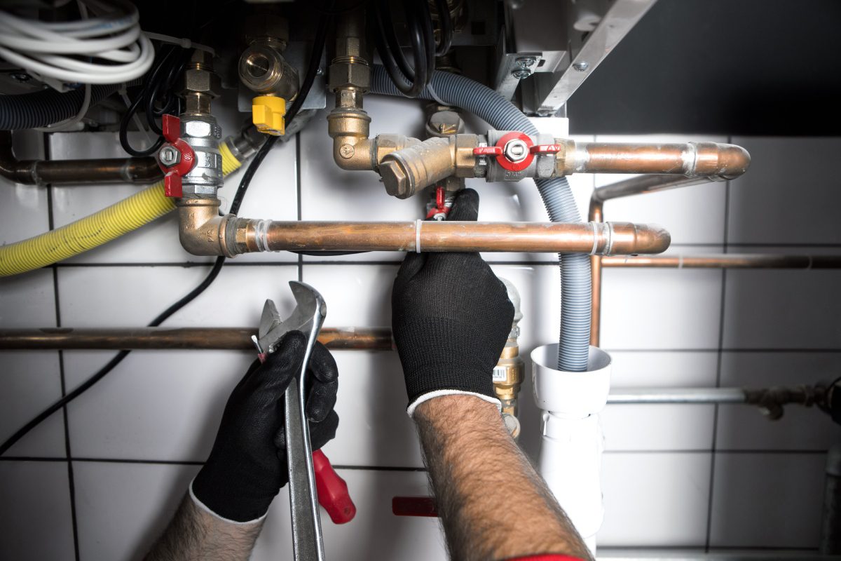 A plumber's hand wearing a glove fixing copper pipes under a sink.